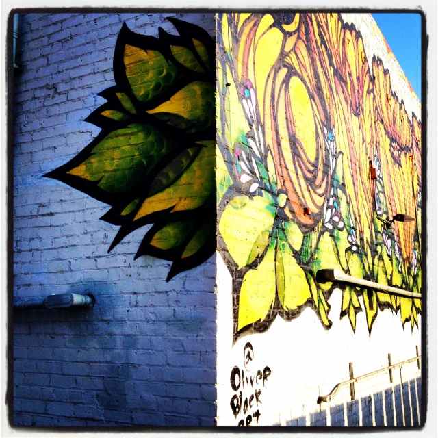 Colorful mural art on a brick wall showing an abstract, nature-inspired design with green leaves, yellow petals, and organic shapes.