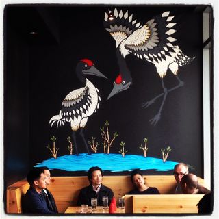 Mural of black and white cranes above group of seated people inside a restaurant with a booth and table setting with dining accessories.