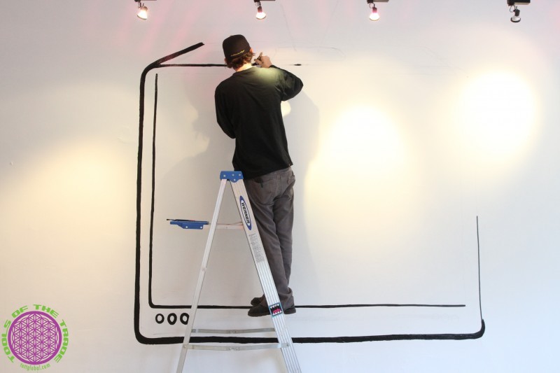 Adult man standing on a ladder and drawing a large, simple digital tablet illustration on a white wall in a brightly lit indoor space.
