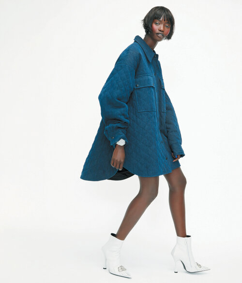 Fashion shot of a Black woman in a blue quilted coat and white boots walking against a white background in a studio environment.