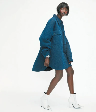 Fashion shot of a Black woman in a blue quilted coat and white boots walking against a white background in a studio environment.