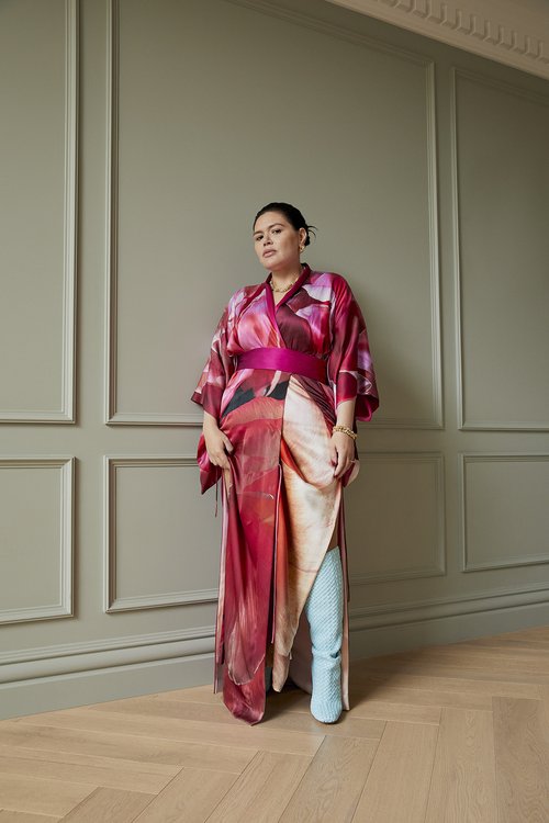 Adult woman posing in an abstract printed kimono dress against a grey panelled wall on a herringbone-patterned wood floor indoors.