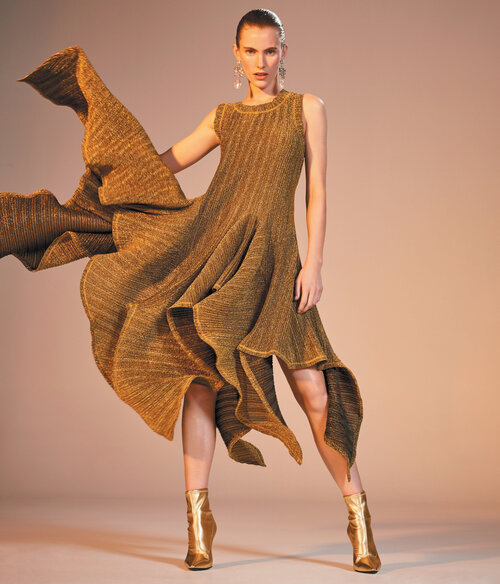 Elegant adult woman in a sleeveless gold dress and matching ankle boots strikes a dynamic pose against a neutral gradient backdrop.