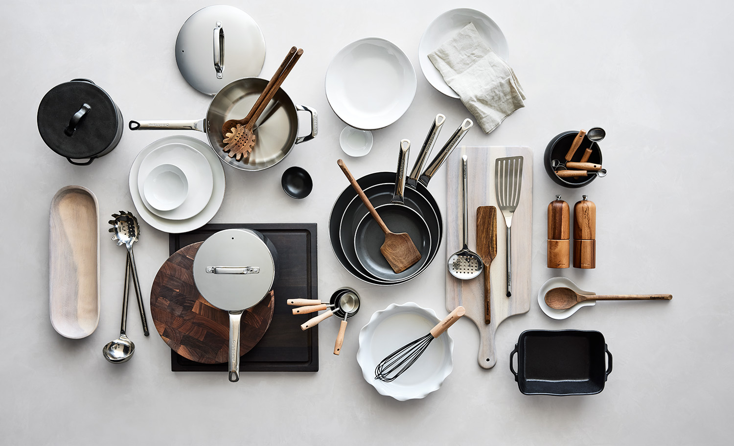Overhead shot of a variety of kitchen utensils, cookware, dishes, and cooking tools arranged on a neutral surface, creating a culinary still life.