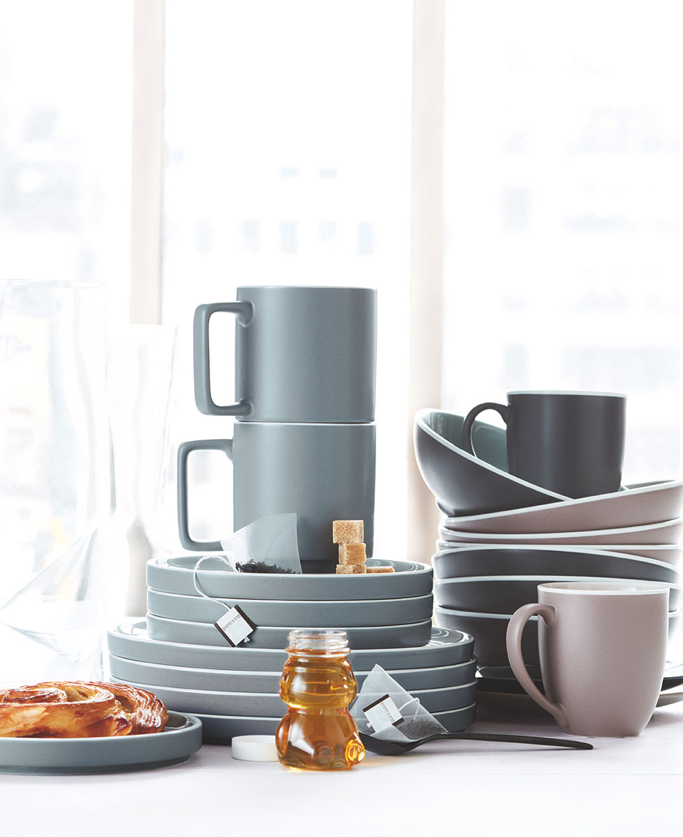 Still life featuring a stack of gray ceramic plates and cups, complemented by dishes, teabags, sugar cubes, honey and a pastry.
