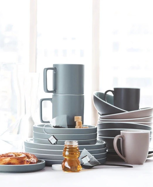 Still life featuring a stack of gray ceramic plates and cups, complemented by dishes, teabags, sugar cubes, honey and a pastry.