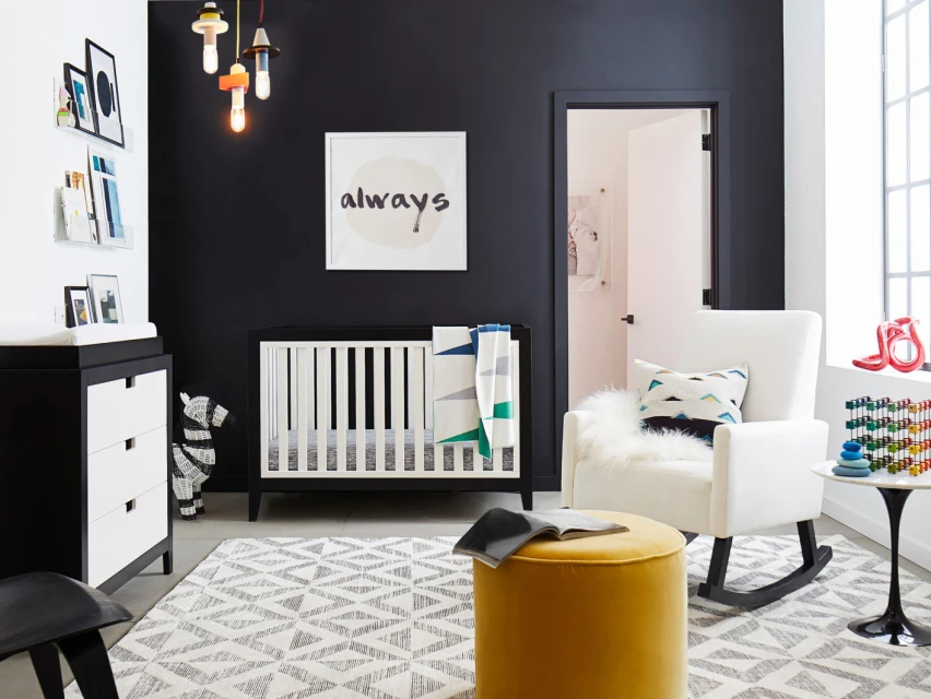 Interior of a modern nursery with a crib, white rocking chair, yellow ottoman, and black accent wall, illuminated by pendant lights.
