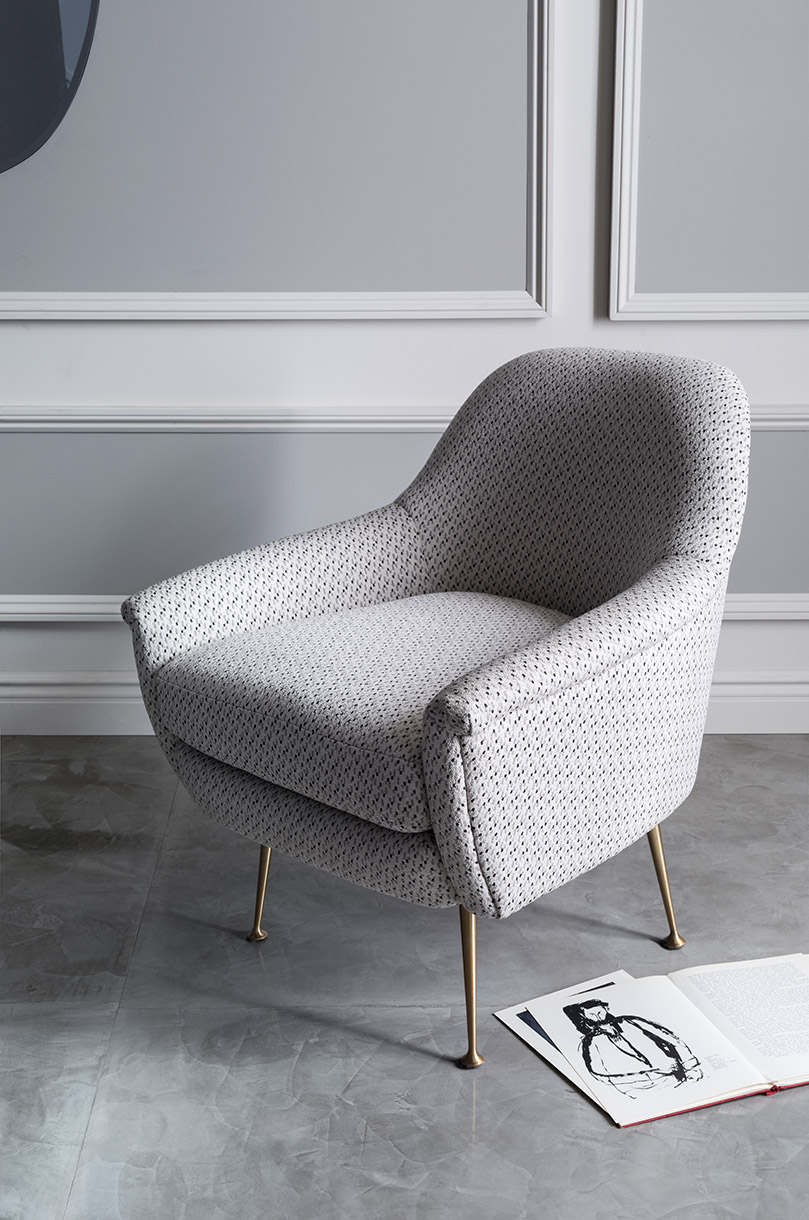 Interior shot of an armchair with patterned fabric, gold legs, placed on a gray floor with a book near a gray wall with molding.