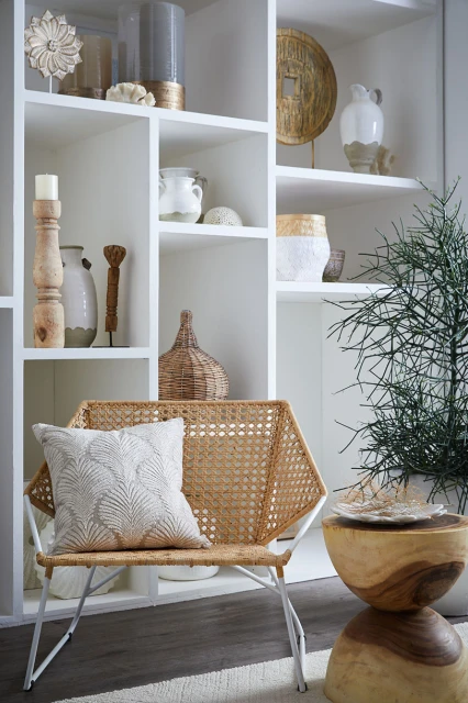 Living room interior featuring a woven chair with a patterned pillow, a wooden side table, and a decorative shelving unit.