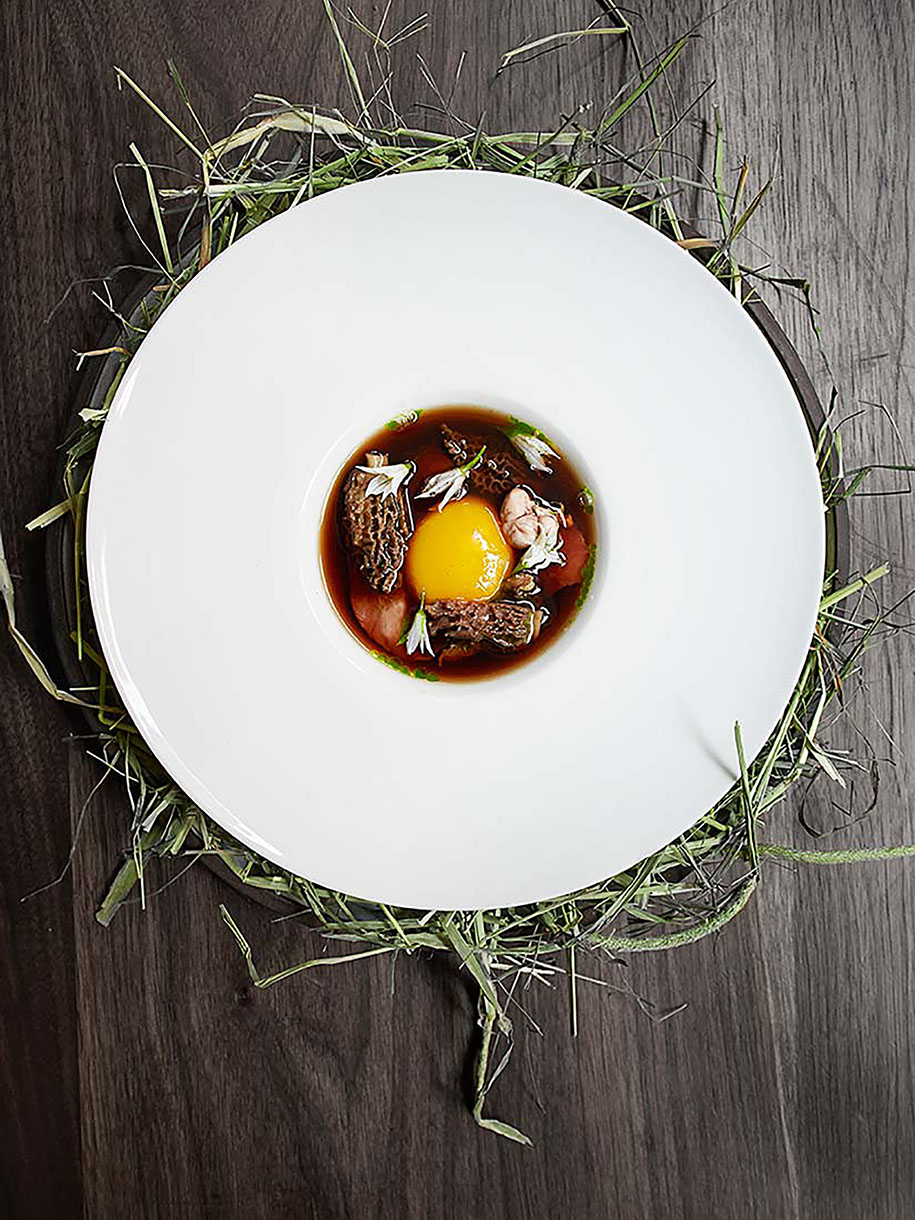 Overhead shot of a white plate with brown broth, mushrooms, a raw egg yolk, and white flowers, set on a wooden table with loose hay.