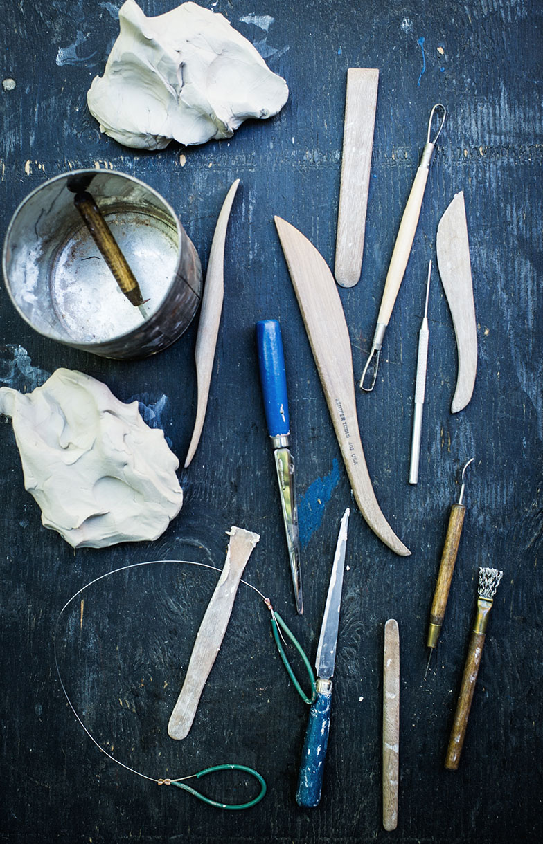 Overhead shot of pottery tools including clay, shaping tools, and wire cutters on a textured, dark blue surface with dim lighting.