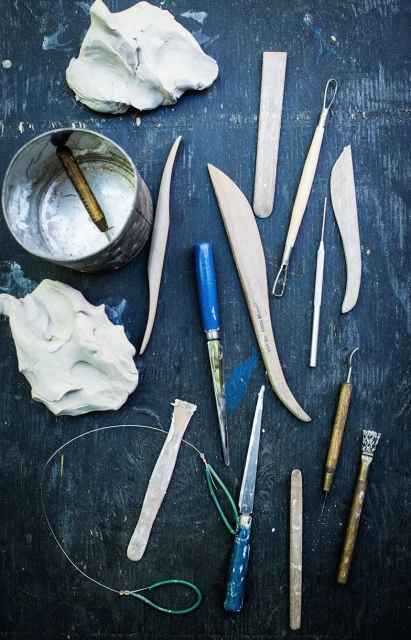 Overhead shot of pottery tools including clay, shaping tools, and wire cutters on a textured, dark blue surface with dim lighting.