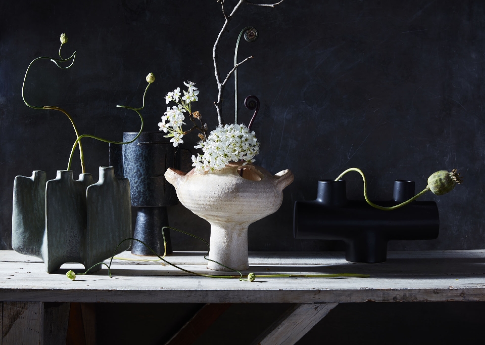 Still life of ceramic vases with flowers and branches on a rustic wooden table against a dark textured background. 