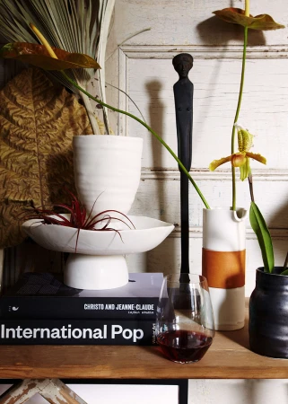 Interior still life featuring a wine glass, vases with flowers, and books, set on a wooden shelf against an off-white door.