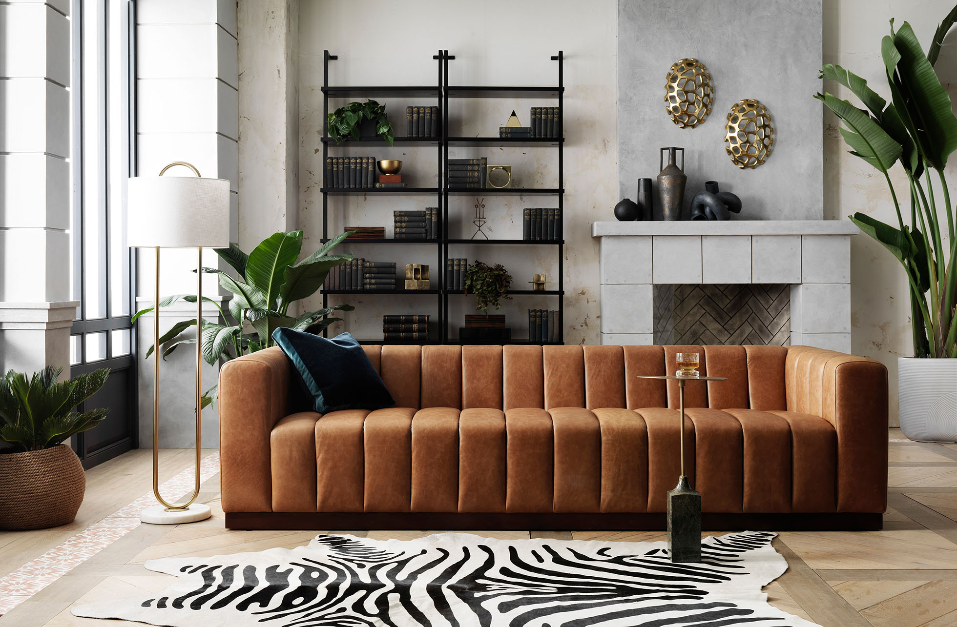 Living room interior featuring a brown leather sofa, black bookshelf, zebra-print rug, floor lamp, fireplace, and potted plants.