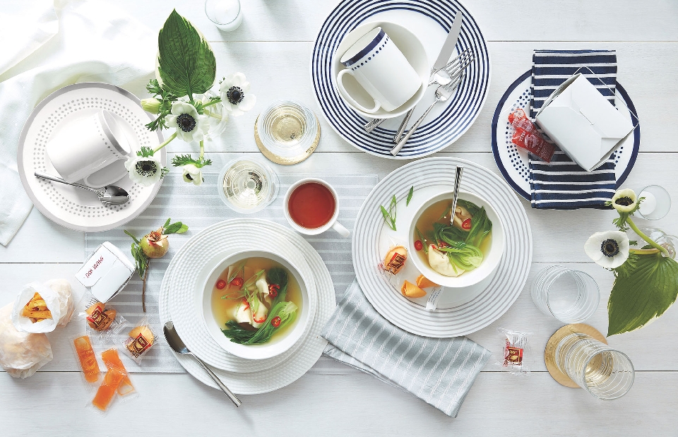 Overhead shot of soup bowls, plates, silverware, and flowers on a white table, conveying a fine dining or food presentation aesthetic.
