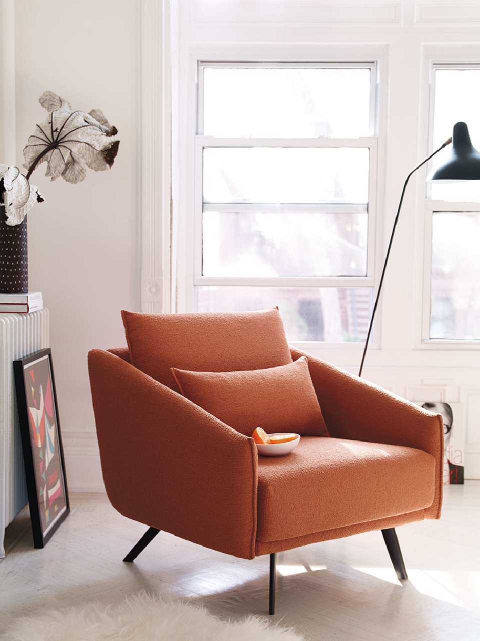 Interior with terracotta-colored modern armchair with black legs and throw pillow, near a window and standing lamp, with decorations.