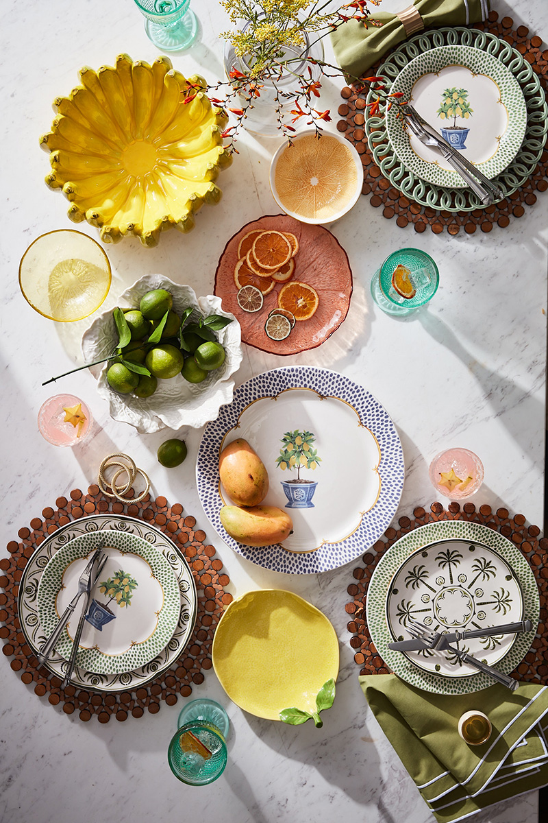Overhead shot of colorful table setting featuring plates with botanical designs, fresh fruit, decorative bowls, and glasses on a white marble surface.