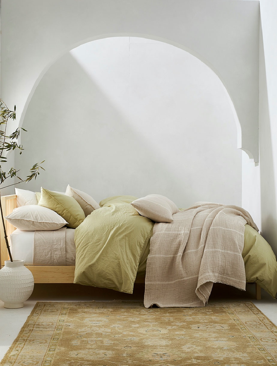 Bedroom interior with a wooden bed frame, green duvet, pillows, a throw blanket, patterned rug, and a white vase with bamboo.