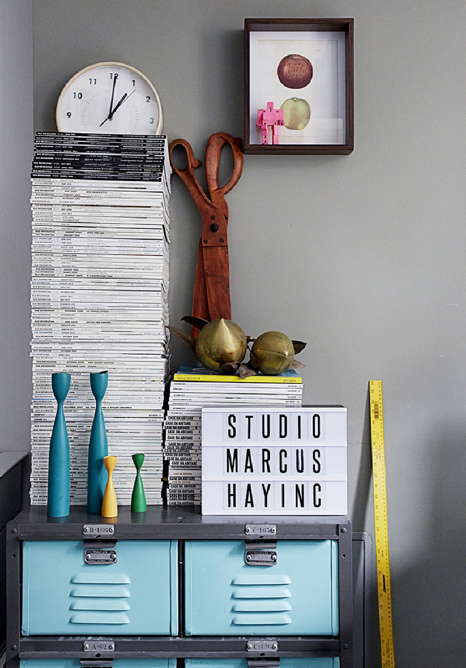 Still life featuring magazines, clock, framed art, scissors, and teal blue lockers, creating a visually rich and textured composition.