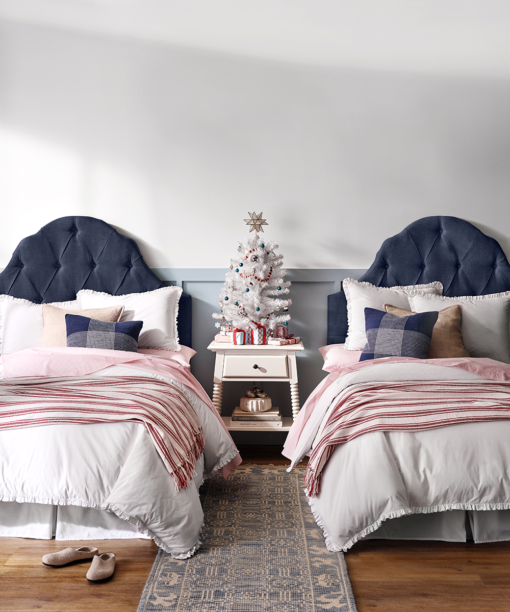 Twin beds in bedroom with blue upholstered headboards, white bedding, red striped throws, and Christmas decor on bedside table.