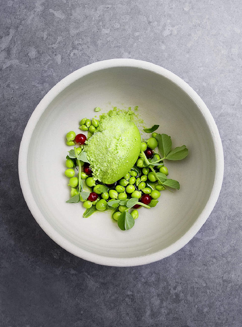 Overhead shot of a modern food dish featuring bright green pea puree, red berries, pea sprouts, and vibrant colors inside a white bowl.