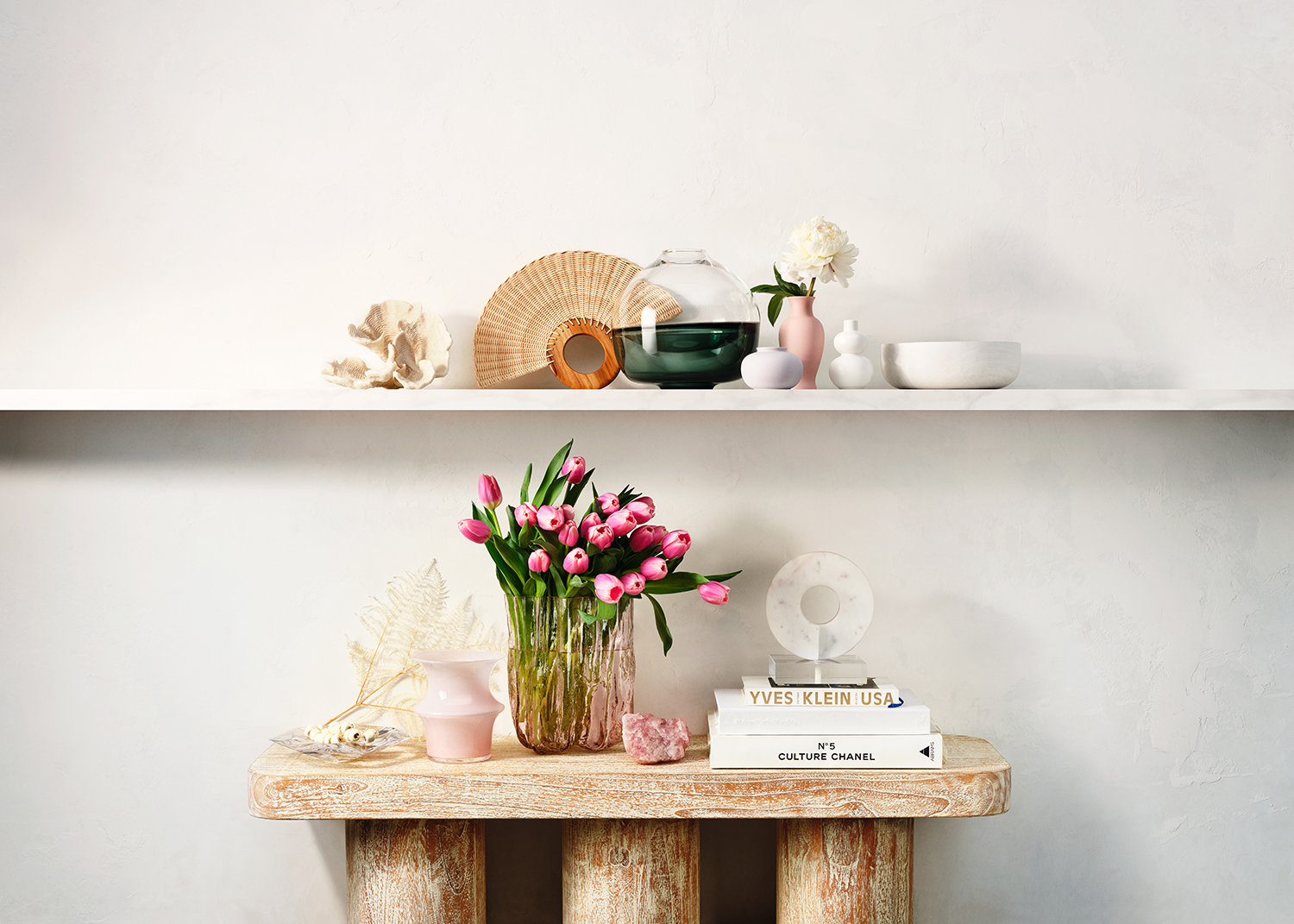 Still life of tulips in a vase, displayed on a wooden table with books and decorative objects against a plain white wall.