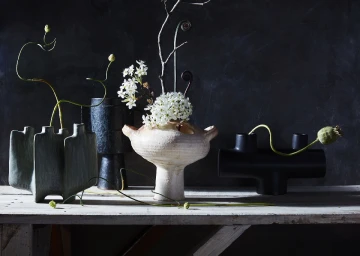 Still life arrangement of ceramic vases filled with dried stems and white flowers on a weathered wooden tabletop against a dark wall
