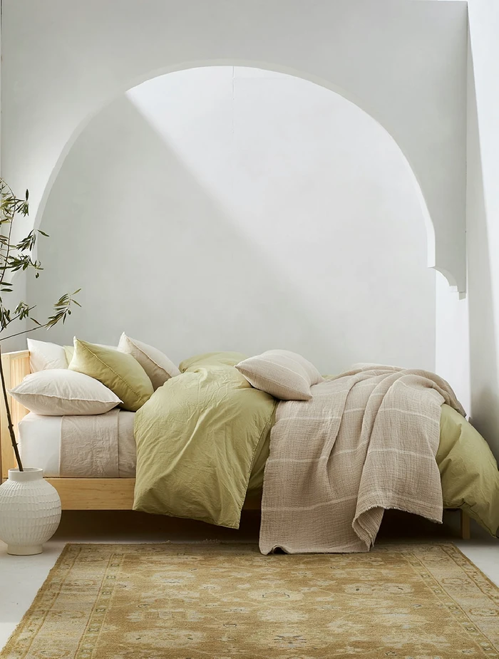 Minimalist bedroom interior styled by Marcus Hay featuring a low wooden bed with sage green bedding and neutral pillows beneath an architectural arch.