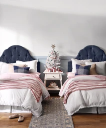 Two twin beds with navy tufted headboards and pink and white bedding separated by a small white nightstand with a decorated mini Christmas tree