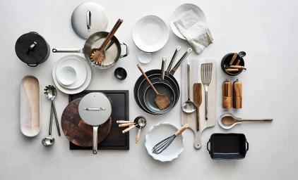 Overhead flat lay of kitchenware including stainless steel pans, wooden utensils, and white plates on a grey surface