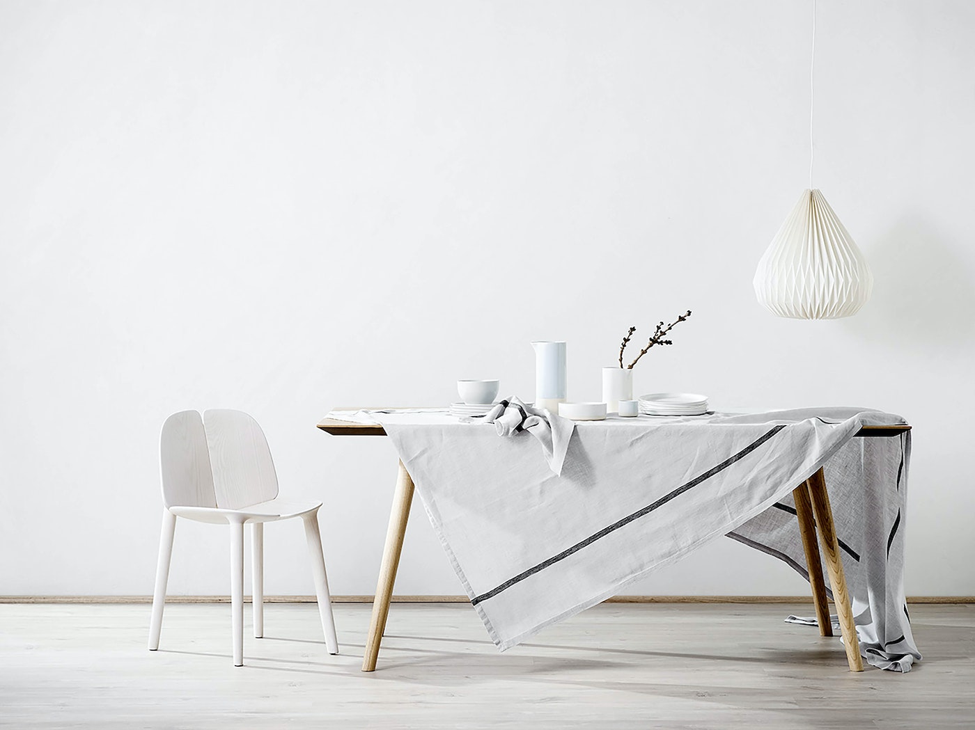 Minimalist dining room interior: white chair, wooden table with grey linen, white dishware, and pendant light against a white wall.