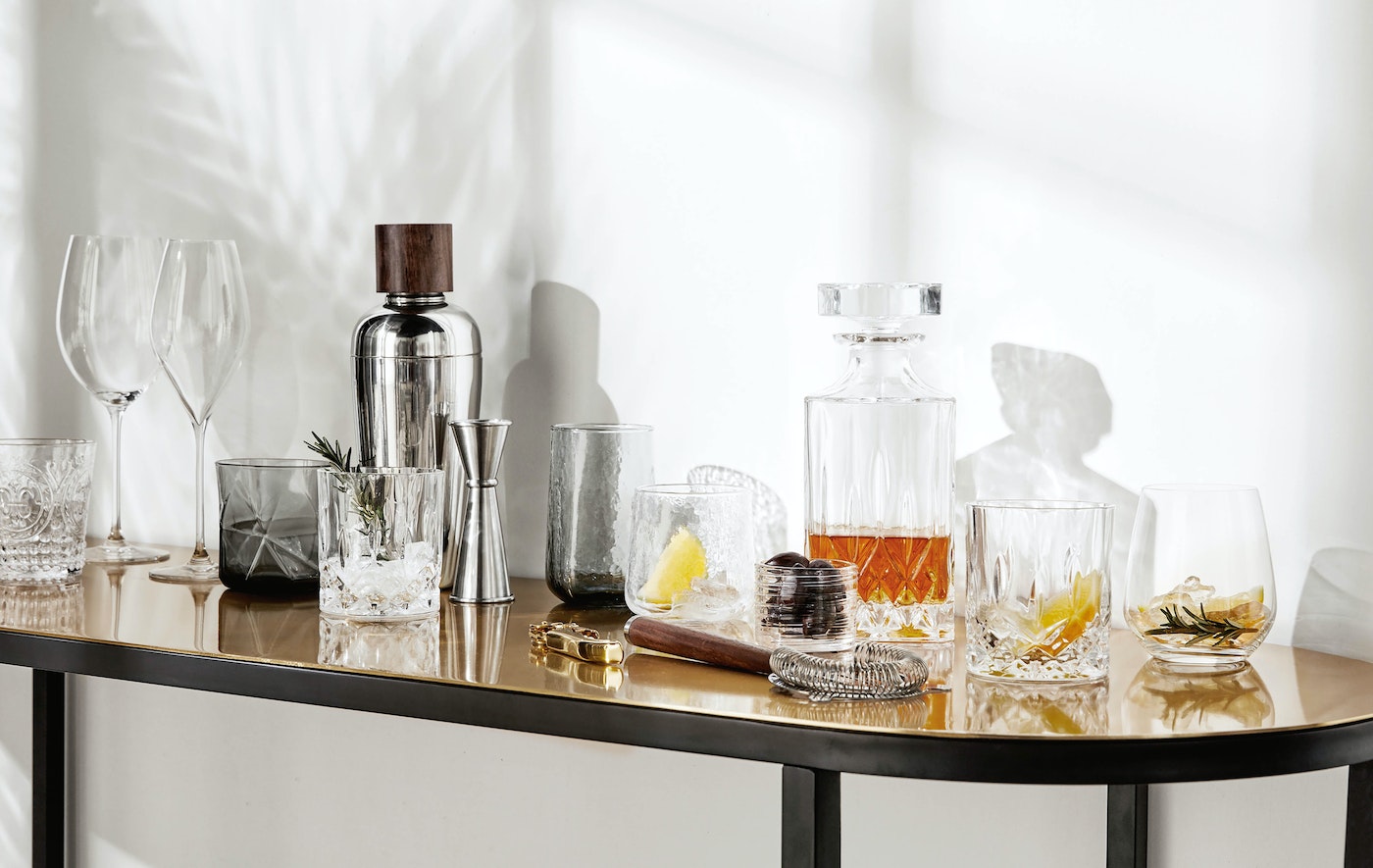 Elegant barware set on a table featuring crystal glassware, cocktail shaker, and various bar tools against a light, airy backdrop.