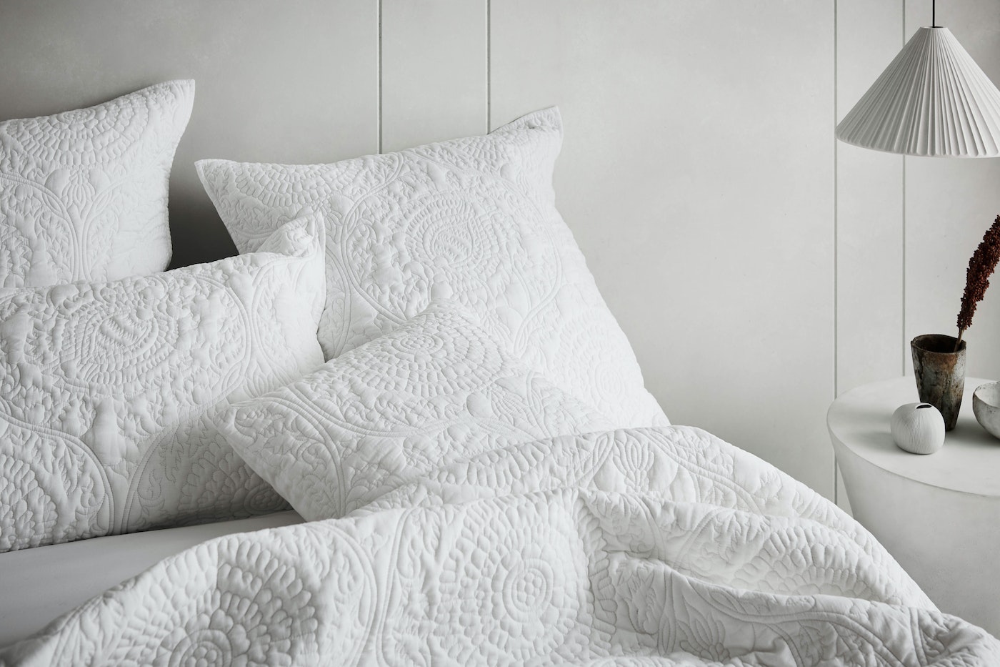 White quilted bedding with decorative pillows arranged on bed in a bright room, accented by a side table and minimalist lighting.