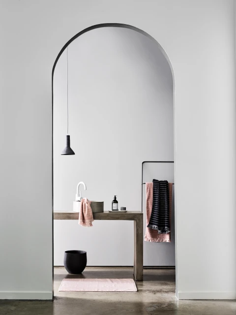 Modern bathroom interior featuring a sink, concrete vanity, black accessories, and a pink bath mat, framed by an arched doorway.