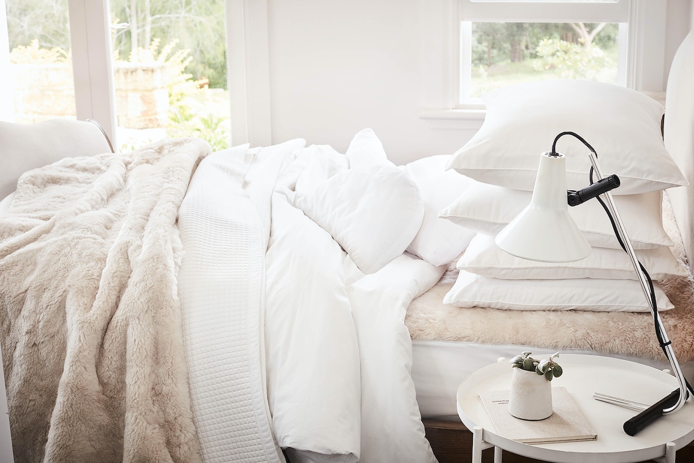 Bedroom interior featuring a bed with white linens and a faux fur throw, illuminated by a modern desk lamp on a small round table.