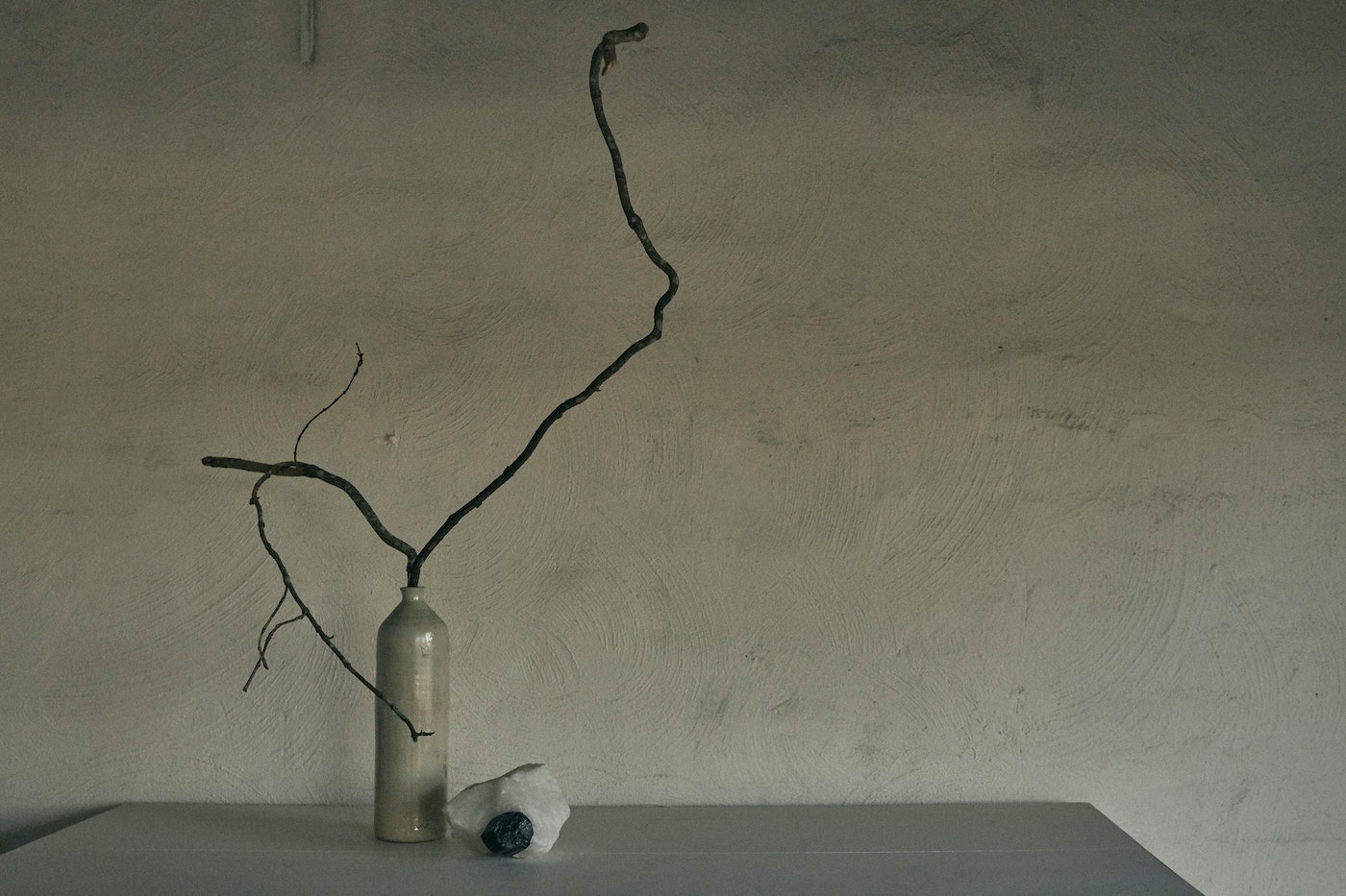 Minimalist composition featuring a dried branch in a ceramic vase alongside a white stone with a dark inclusion on a gray surface.
