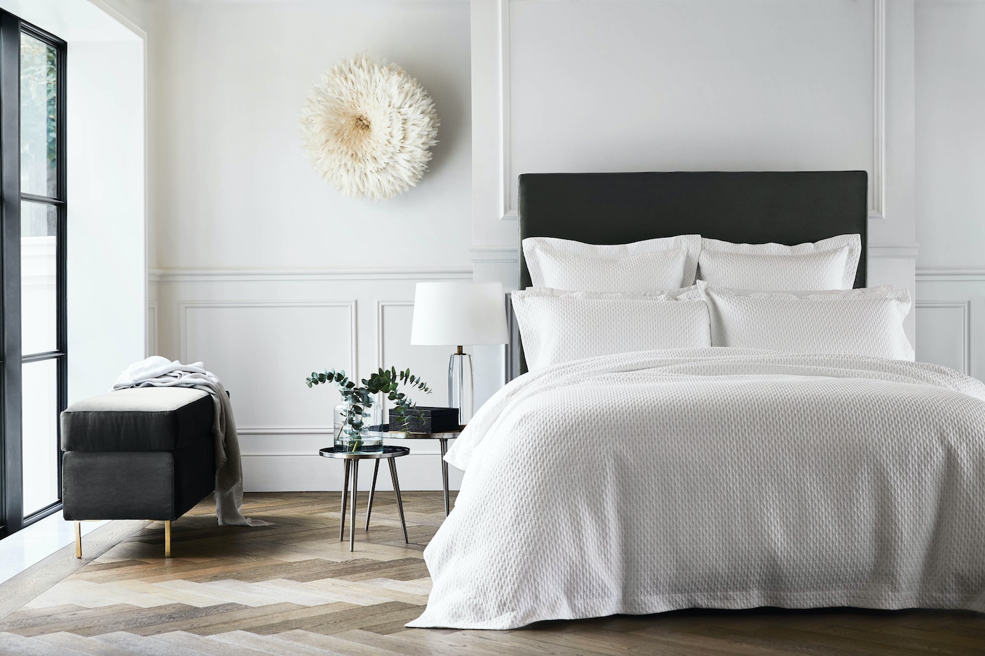Bedroom interior showcasing a bed with white textured linens, pillows, and a dark headboard, complemented by a bench and wooden floor.