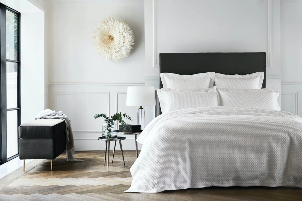 Bedroom interior showcasing a bed with white textured linens, pillows, and a dark headboard, complemented by a bench and wooden floor.