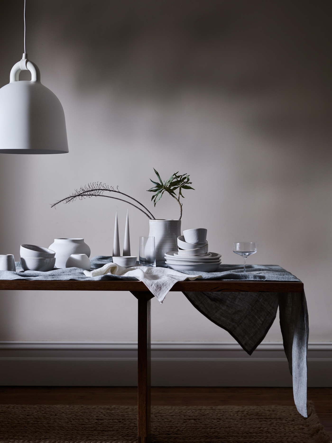 Still life indoor scene featuring ceramic tableware on a wooden table with a linen cloth, beneath a hanging lamp, against a gray wall.
