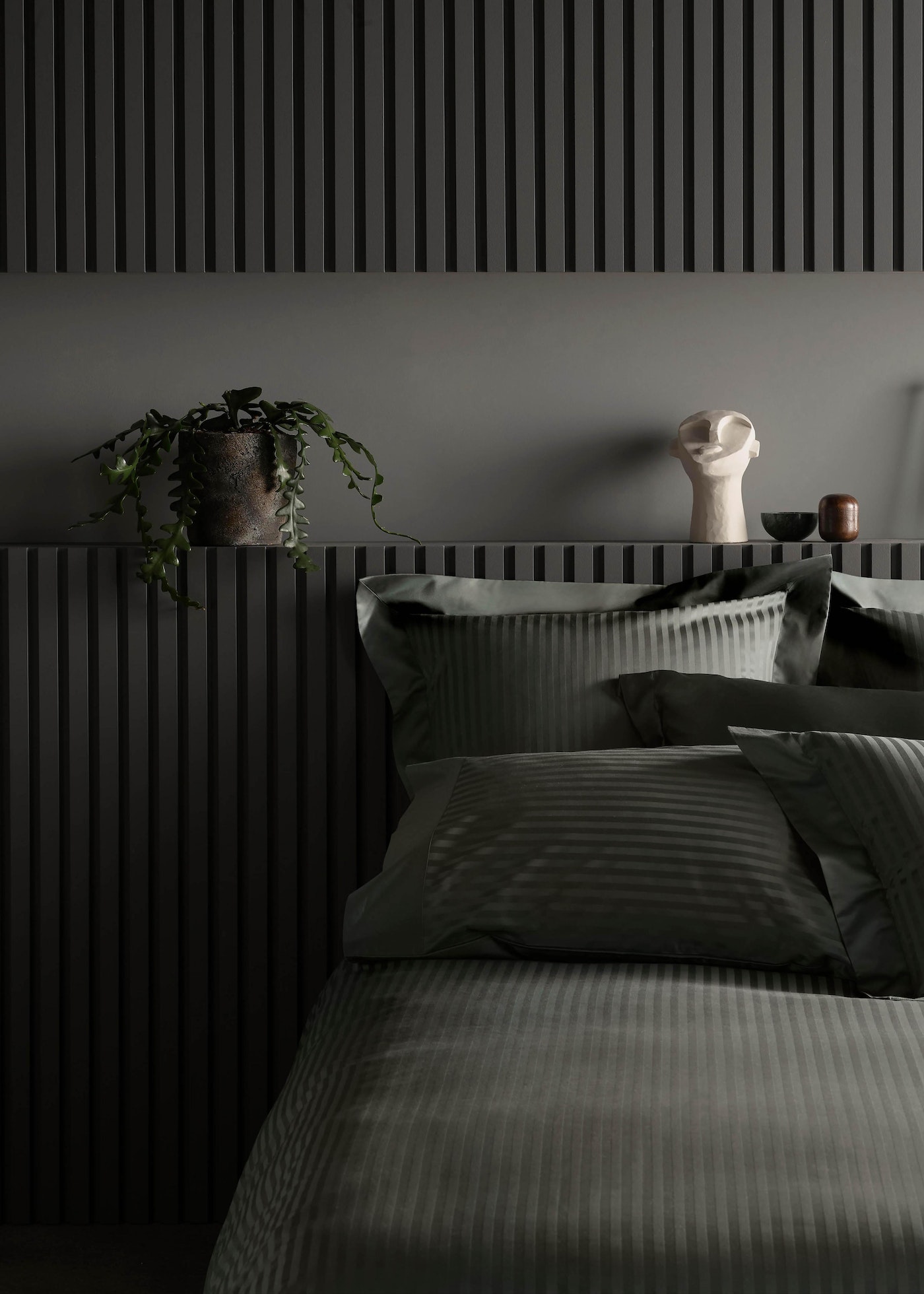 Bedroom interior with grey striped bedding, grey slatted wall paneling, potted plant, and decorative white head sculpture on shelf.
