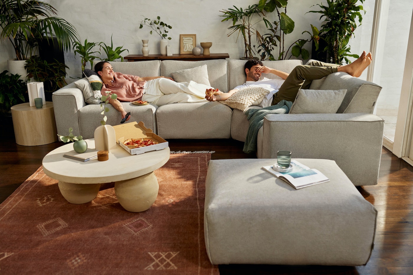 Couple relaxes on a sectional sofa in a bright living room with modern decor, pizza, plants, and a brown rug. Leisure and comfort.