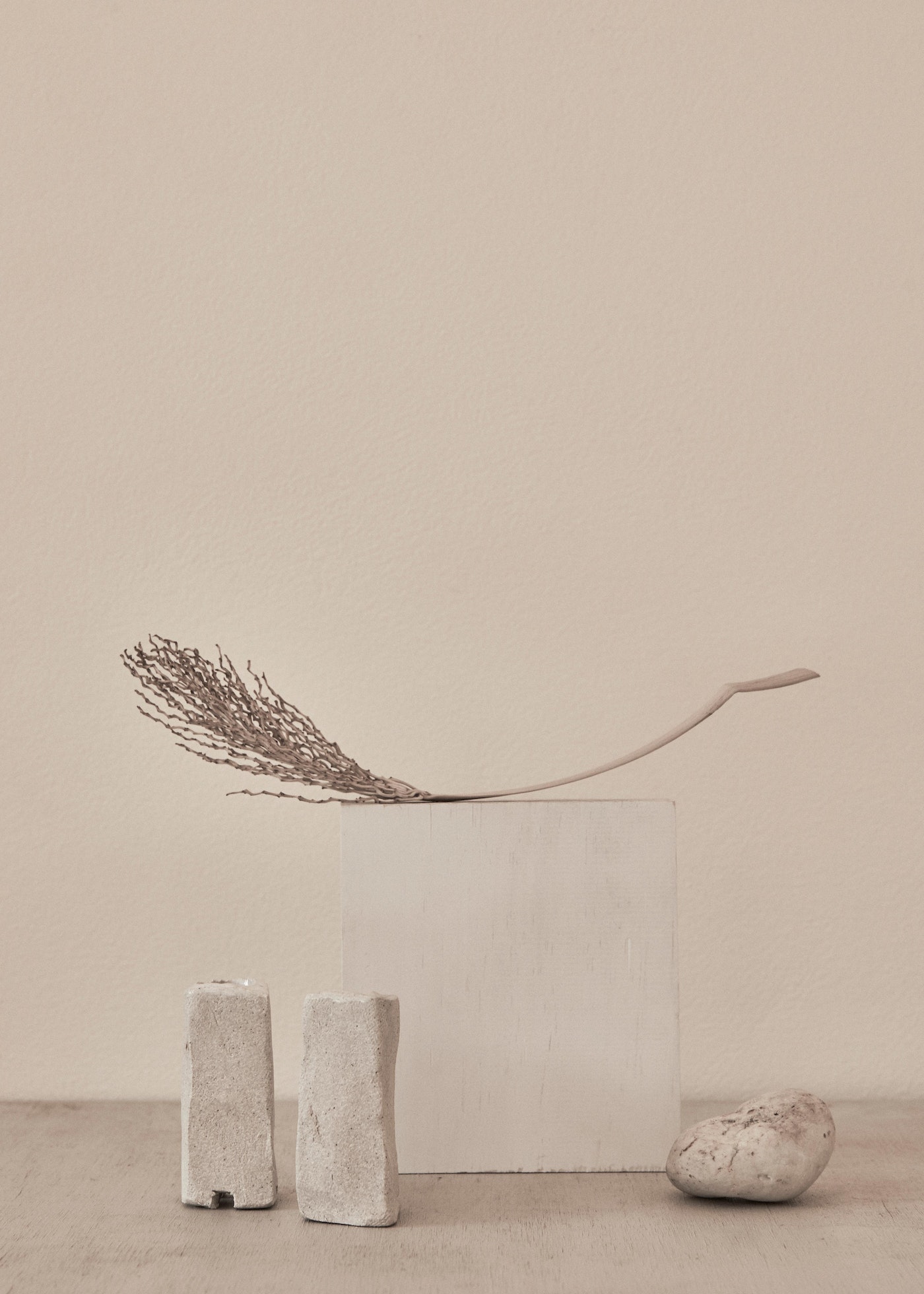 Neutral minimalist product photography of a feather laying on a concrete cube, flanked by two stone-like candle holders and a stone.