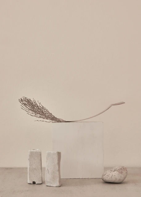 Neutral minimalist product photography of a feather laying on a concrete cube, flanked by two stone-like candle holders and a stone.