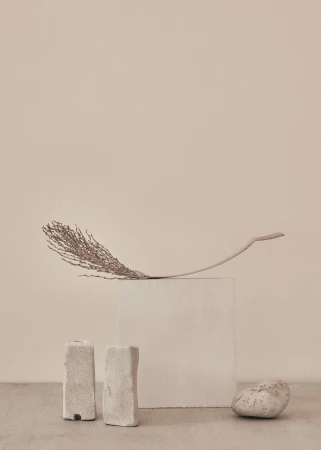 Neutral minimalist product photography of a feather laying on a concrete cube, flanked by two stone-like candle holders and a stone.