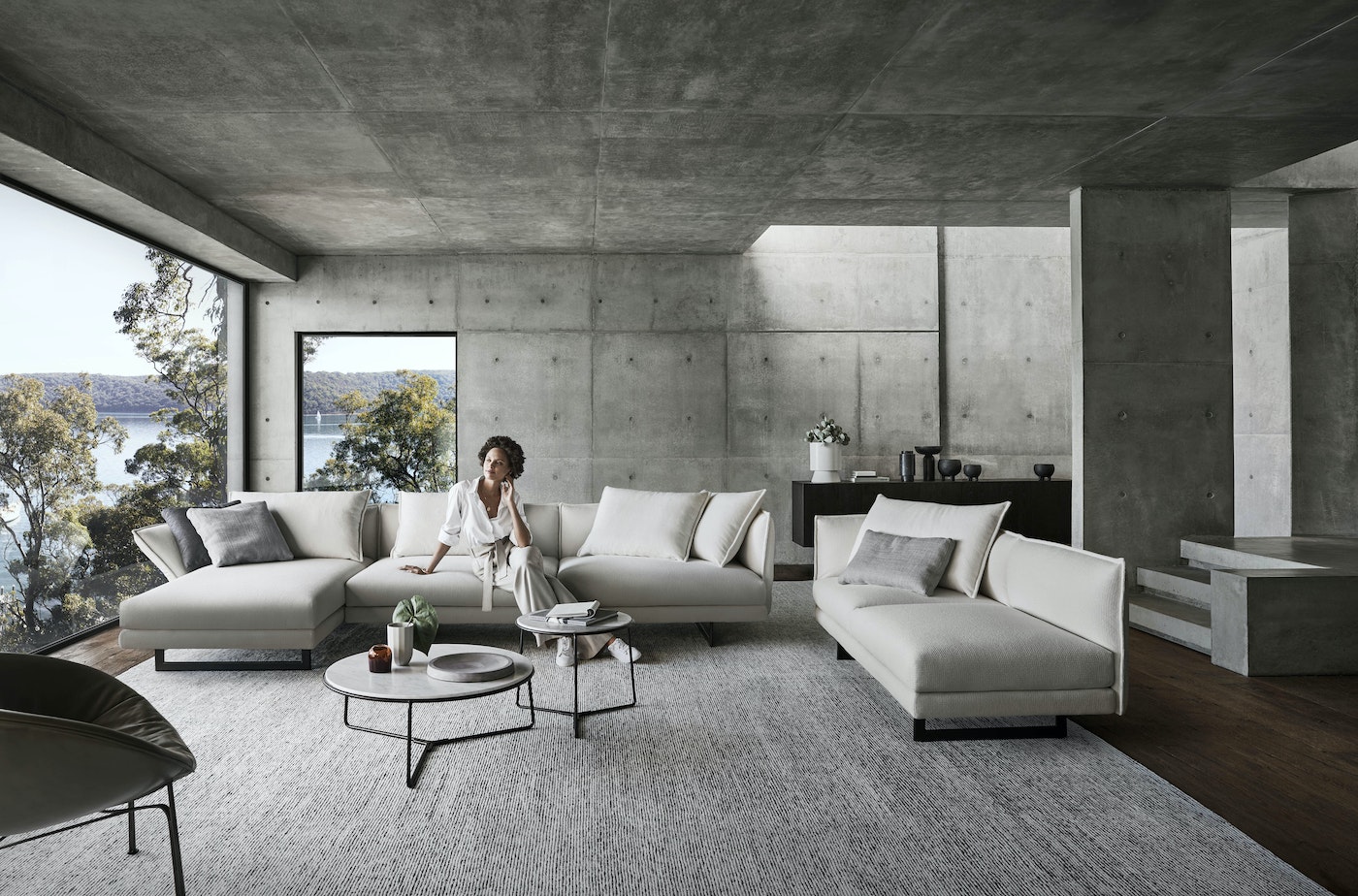 Contemporary living room with a woman seated on a sectional sofa, complemented by neutral tones, concrete walls, and natural light through large windows.