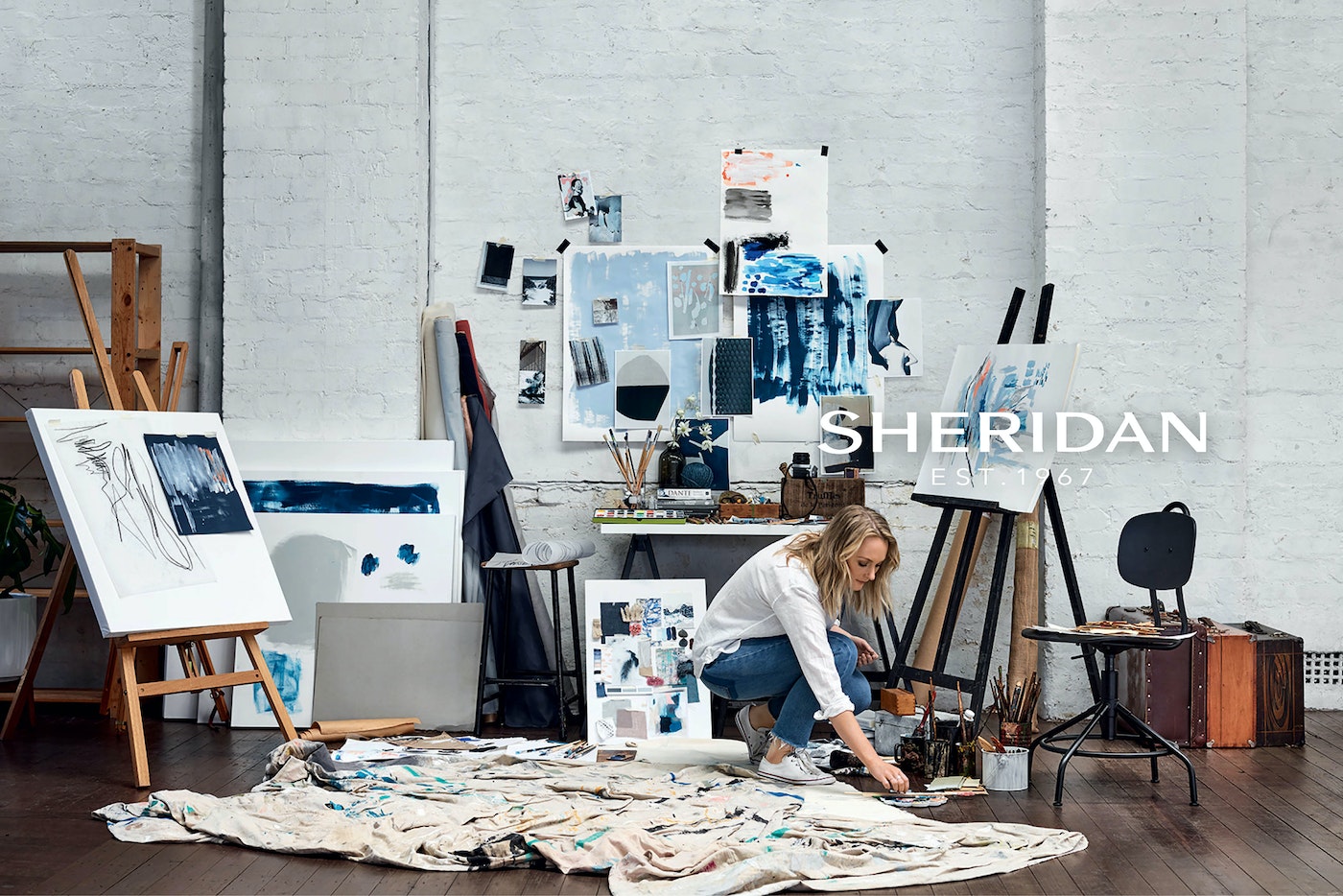 Woman artist in studio working on abstract paintings surrounded by art supplies, easel, and color palettes in daylight.