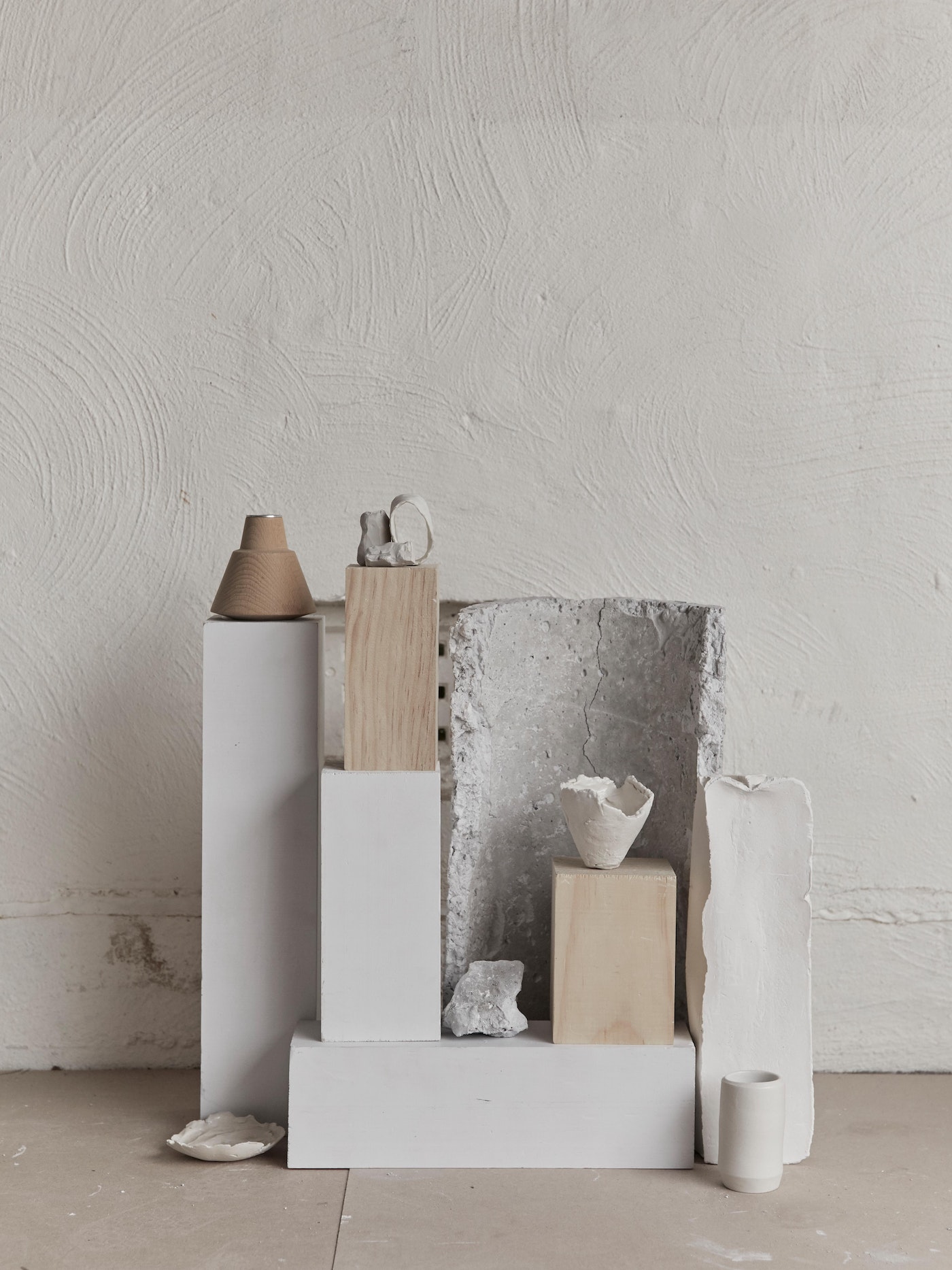Still life arrangement of ceramic art pieces and geometric pedestals against a textured white wall in a minimalist composition.