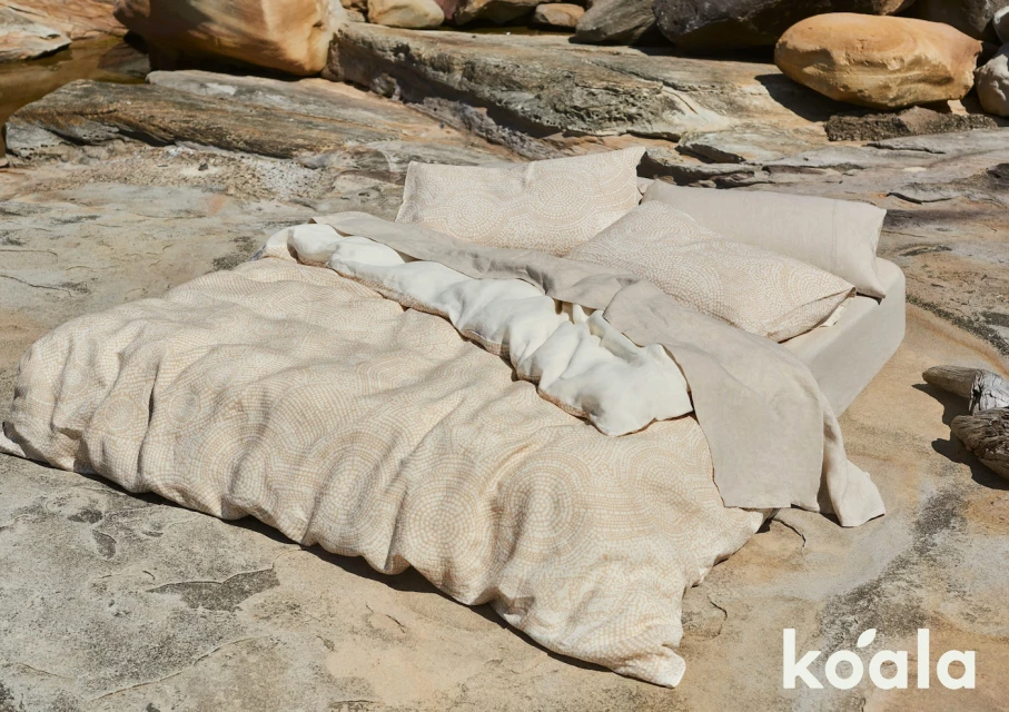 Beige patterned bedding set on a bed placed outdoors on a rocky surface near large rocks. The bedding includes pillows and duvet.