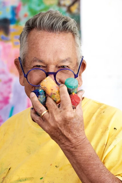 Elderly male artist with paint-covered hands holds paint pots in an indoor studio with an abstract painting visible in the background.